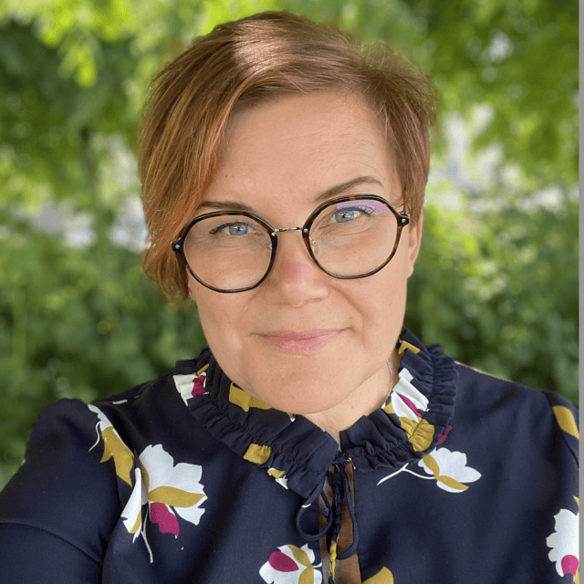 Riikka Marttinen katsoo suoraan kameraan. Hänellä on silmälasit ja punertavat lyhyet hiukset.