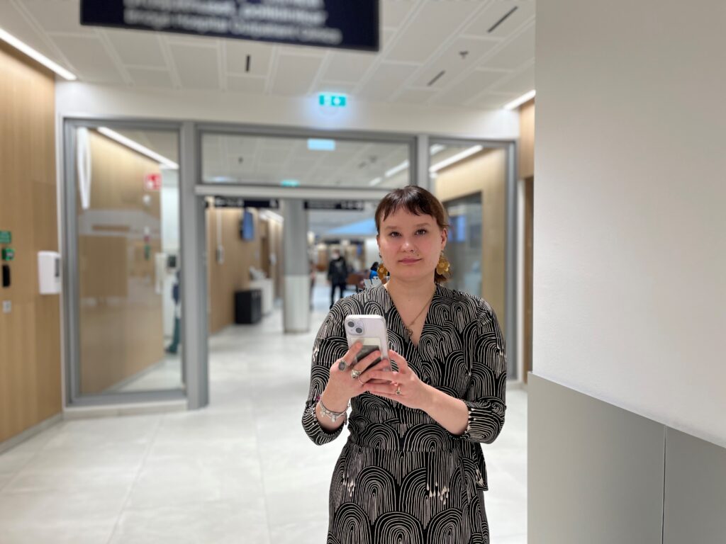 Nea Mäkinen seisoo taustallaan sairaalan käytävä,pitää molemmilla käsillään kiinni kännykästään ja katsoo suoraan kameraan.